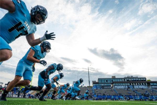 A line of football players run across a football field. They are seen from the ground looking up.