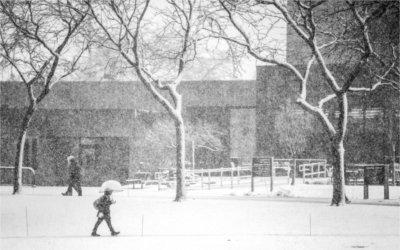 People walk across a snowy campus. One person is carrying an umbrella.